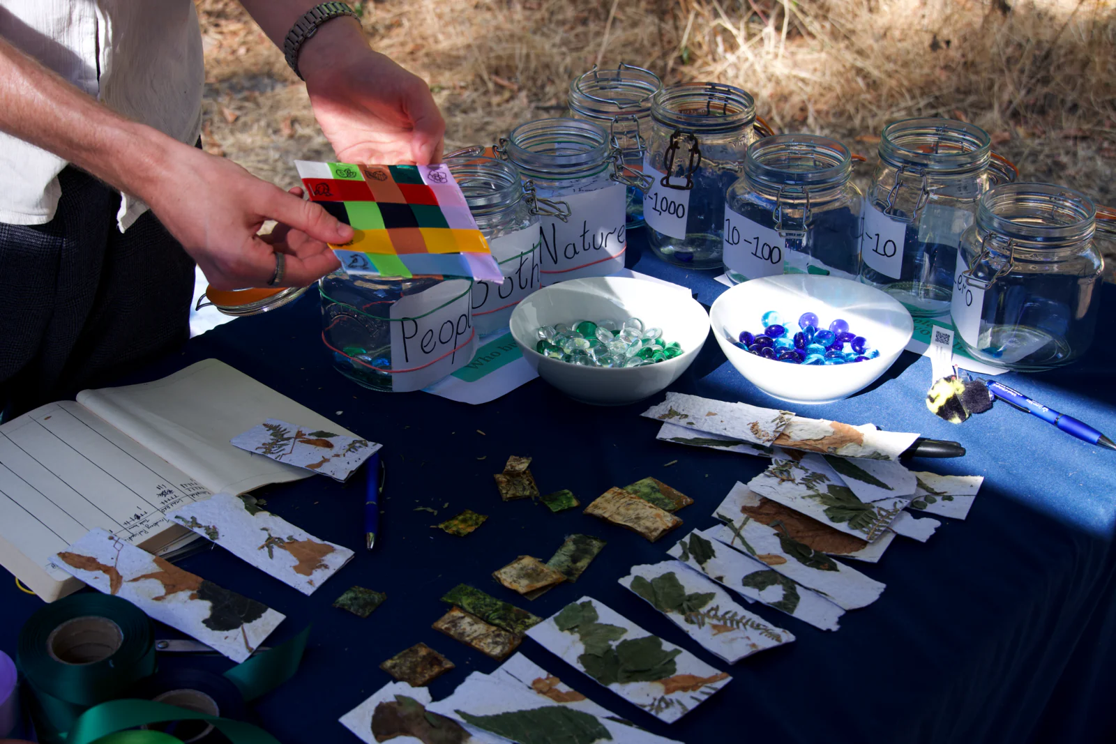 The stall at the fair: a hand holds up a small woven Fabric of Life diagram of coloured ribbons above a table of glass voting jars, bowls of glass beads and squares of handmade leaf paper