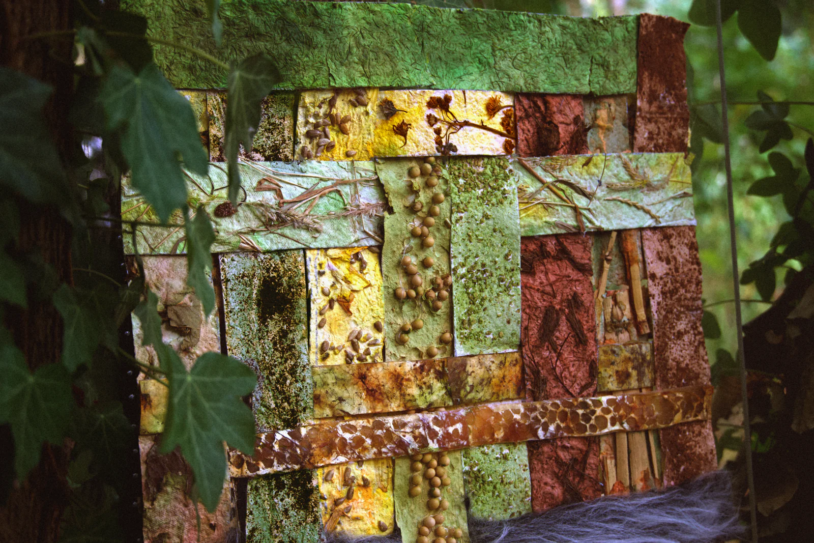 Close-up of the installation: strips of leaf-matrix paper studded with seeds and pressed plants, woven with mycelium textile and a tuft of grey fur