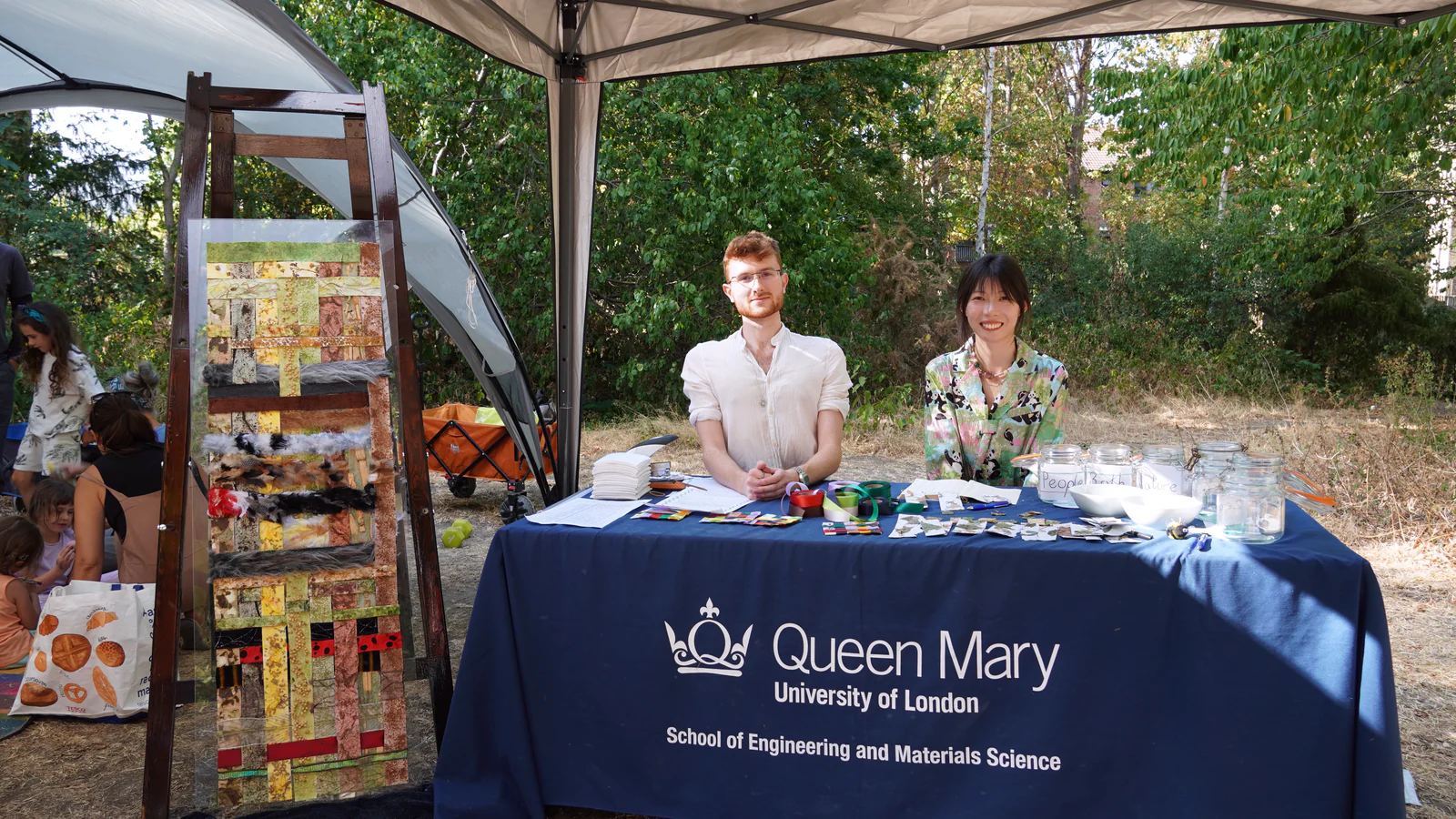 Kristian and Qi sitting at the Queen Mary stall under a gazebo at the summer fair, the installation standing on a wooden easel beside them